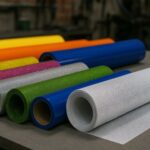 Rollos de vinil textil glitter en varios colores, vinil PVC en tonos sólidos y un rollo de vinil microperforado sobre una mesa de trabajo en un taller de impresión, con fondo industrial desenfocado.