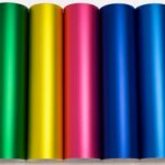 Rollos de vinil automotriz metálico en colores vibrantes como morado, verde, amarillo, rosa, azul y rojo, alineados horizontalmente sobre fondo blanco con cuadrícula, ideales para car wrapping.