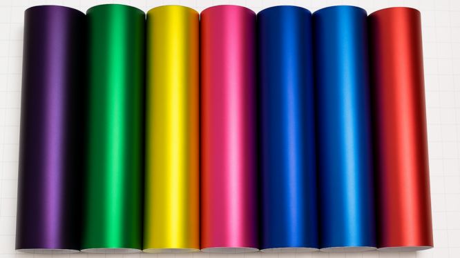 Rollos de vinil automotriz metálico en colores vibrantes como morado, verde, amarillo, rosa, azul y rojo, alineados horizontalmente sobre fondo blanco con cuadrícula, ideales para car wrapping.